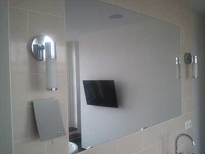 Een rechhoekige make up spiegel, in een grijs betegelde badkamer, met badkamerlampen aan de zijkant van de spiegel en een zilveren kraan