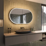 Deze fraaie ovale spiegelkast van aluminium is van alle gemakken voorzien, zoals: geïntegreerde verlichting, spiegelverwarming in de deuren, 2 geaarde stopcontacten, soft-close scharnieren en traploos verstelbare glazen plankjes